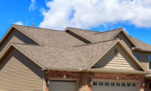 Close-up view of a house with a gable roof covered in light brown shingles against a blue sky.