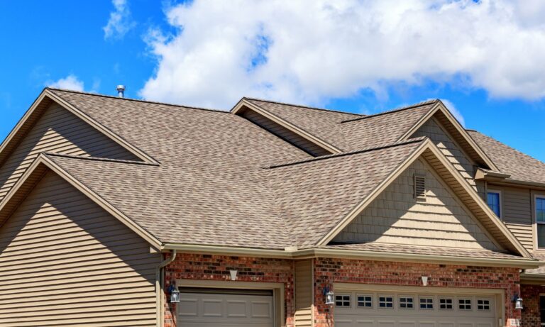 Close-up view of a house with a gable roof covered in light brown shingles against a blue sky.